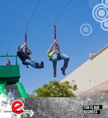 Corporate team building event 2 zip lines 2 men gliding down a zip line, over a green tree while attending a fun corporate event. there's a beautiful clear blue sky behind them.