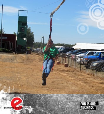 Mobile Zip Wire Hire from Skyfall A team member of a mobile zip wire hire company testing the line after the main tower had been erected. The man is gliding over a sandy area with a line of cars to the right of the image.